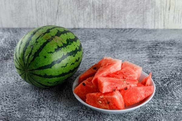 On a stone counter lies a whole watermelon with watermelon slices stacked on a white plate beside it.
