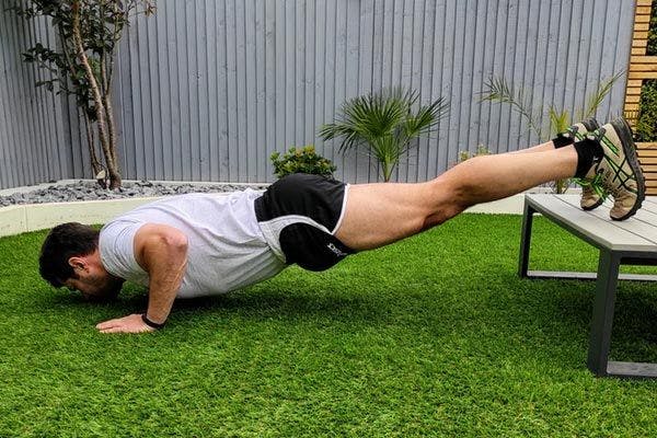 A muscular man with his feet raised on an outdoor bench performing decline push-ups.
