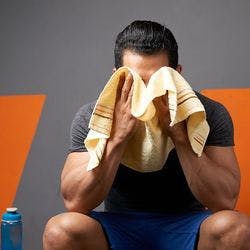 A muscular man resting between sets on a gym bench while wiping the sweat off his face with his towel. 