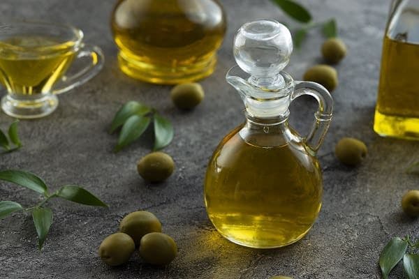 A glass pourer full of olive oil with olives scattered around the base on a stone bench top.